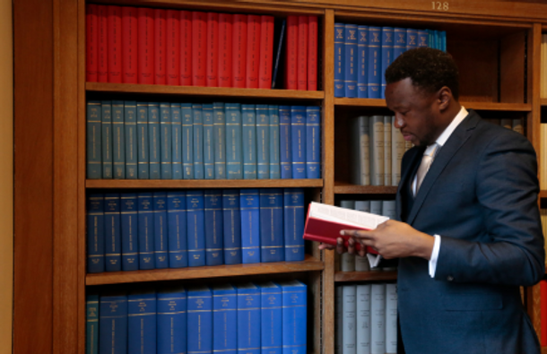 Man holding a red book while standing in front of a bookshelf filled with books.